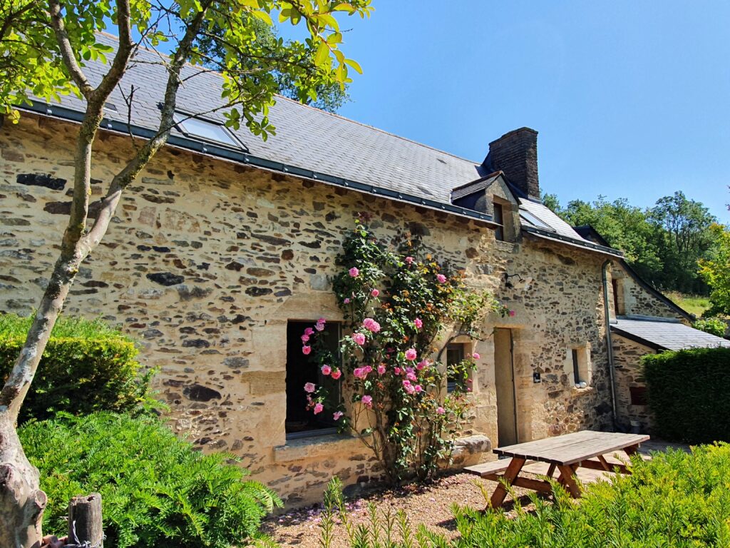 Bonnezeaux: Stone cottage with climbing roses and a wooden picnic table in a garden setting under a clear blue sky.