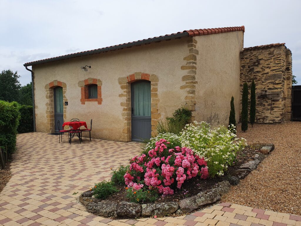 Chinon - A rustic stone building with red trim, featuring a patio with a small table and chairs, and a garden with pink and white flowers in front.