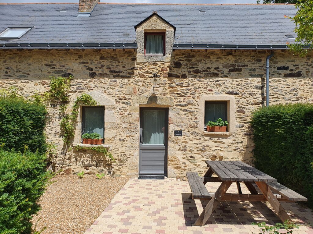 Stone cottage with a slate roof, two windows with potted plants, a central door, and a wooden picnic table on a paved patio.