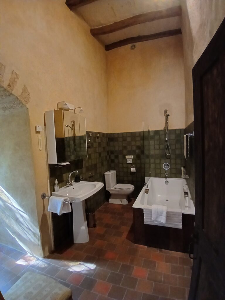 Rustic bathroom with a bathtub, sink, and toilet; tiled walls and floor, exposed wooden beams on the ceiling.