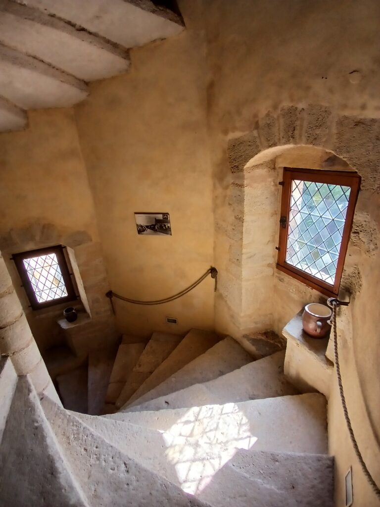 Stone spiral staircase with small windows featuring diamond-patterned glass, lit by sunlight.